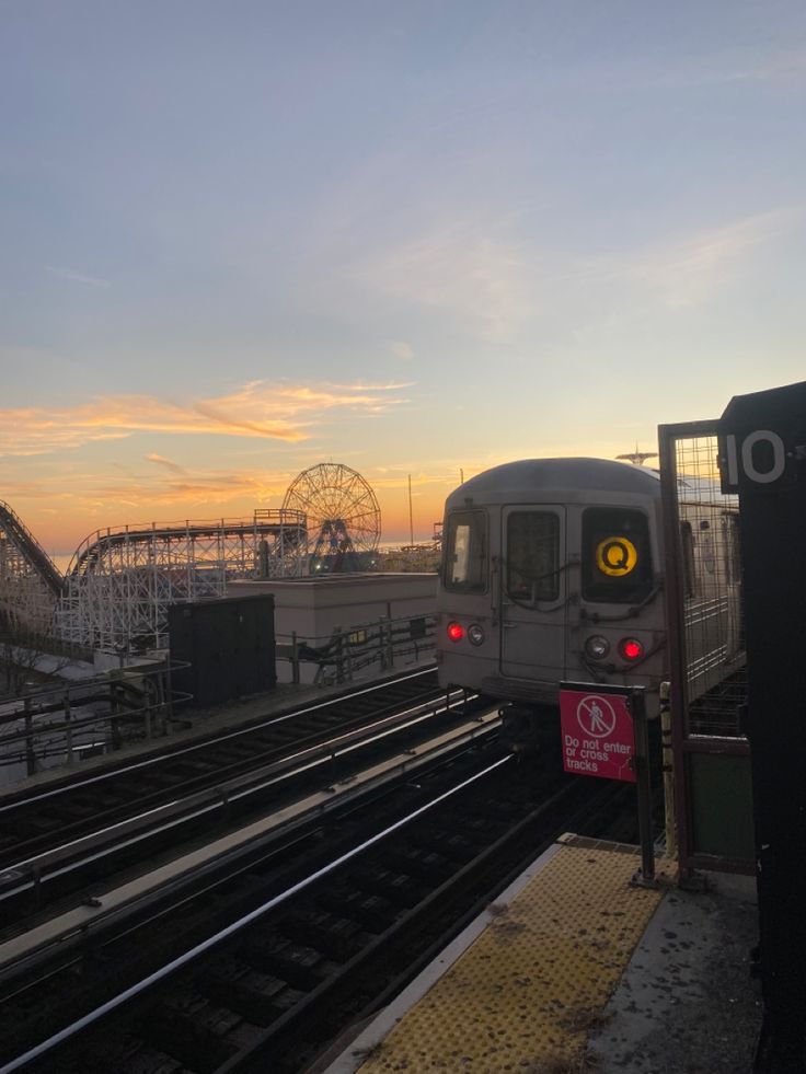 a silver train traveling down tracks next to a roller coaster at dusk with the sun setting in the background