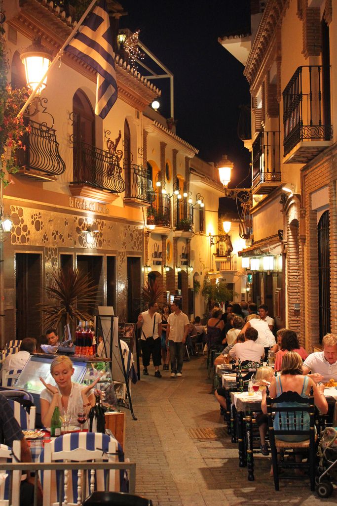 people are sitting at tables in an alleyway with lights on the buildings behind them