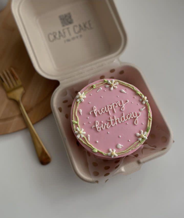 a pink birthday cake sitting in a container next to a fork and spoon on top of a table