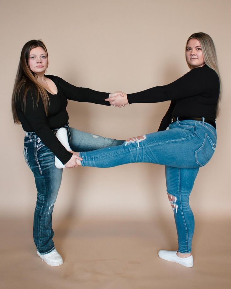 two women in jeans and black shirts are holding each other's hands while standing on one leg