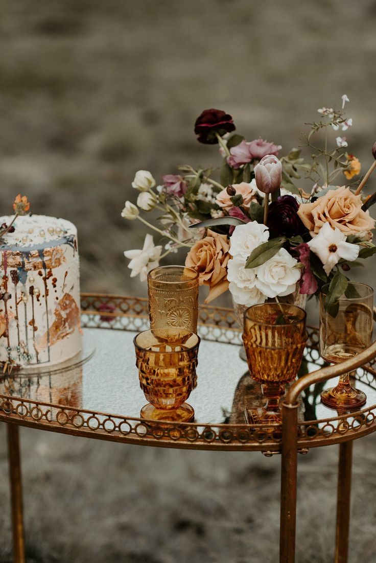 a table topped with two gold vases filled with flowers next to a cake covered in frosting