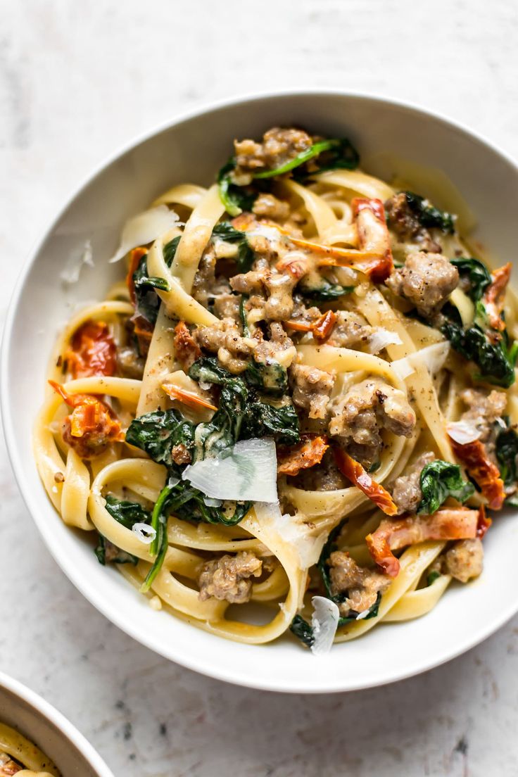 a white bowl filled with pasta, meat and spinach on top of a table