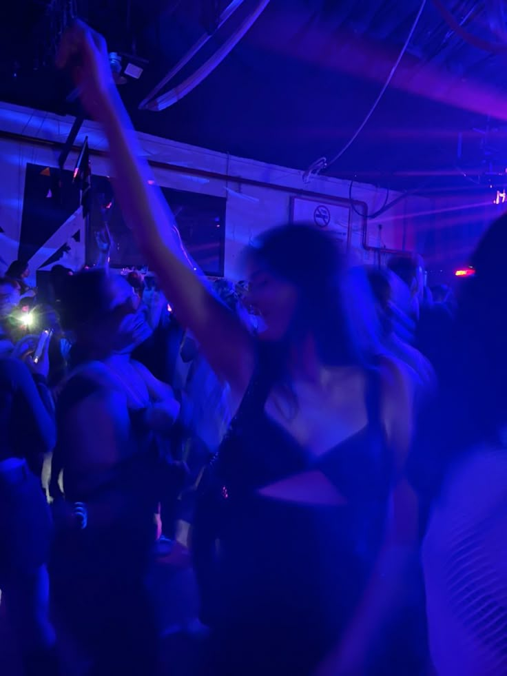 a group of people dancing in a dark room with blue lights on the ceiling and one woman holding her arm up