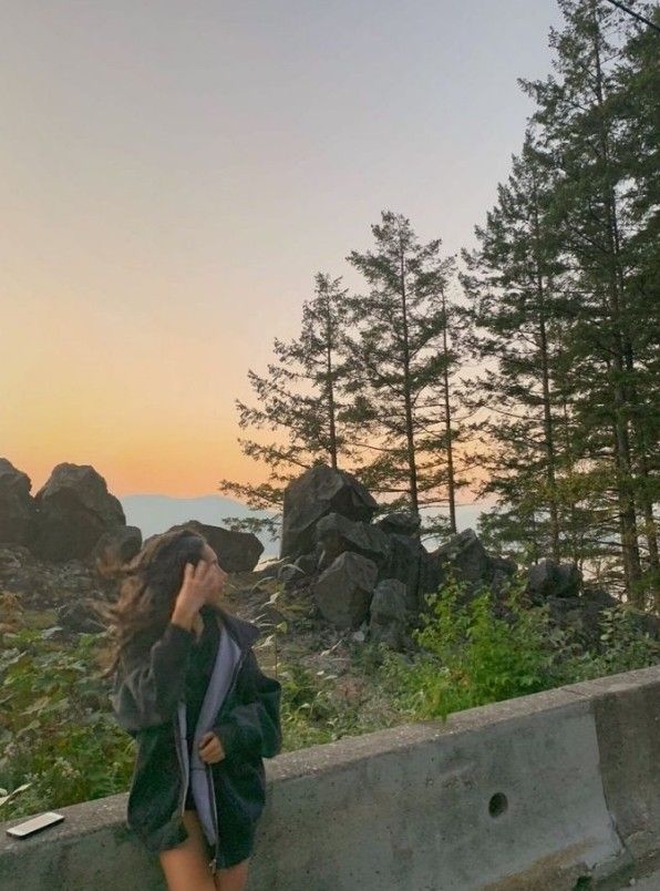 a woman standing on the side of a road next to some trees and water with her hair blowing in the wind