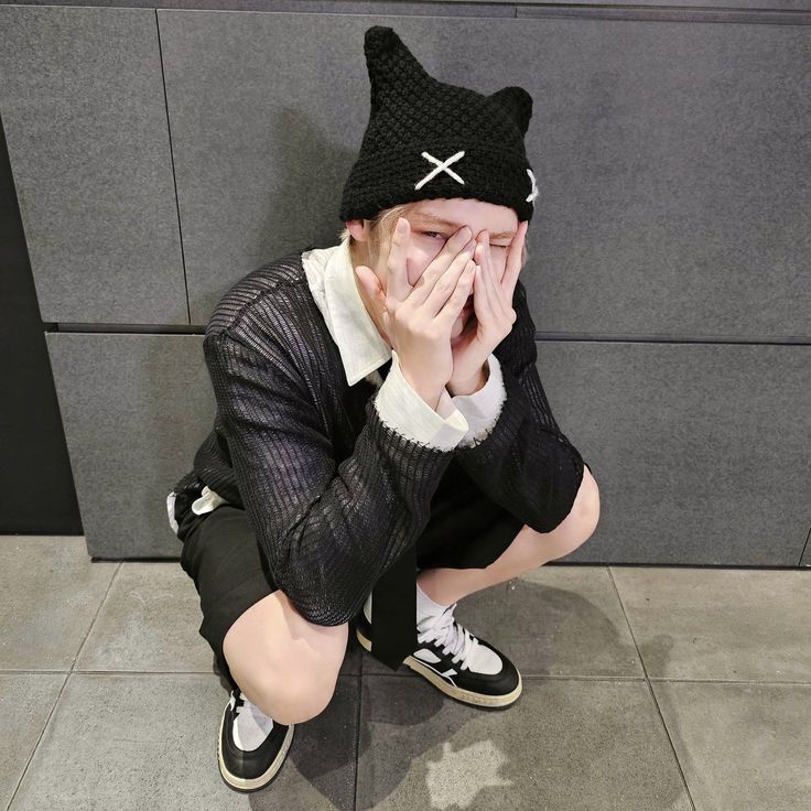 a young man sitting on the floor covering his face with his hands while wearing a black cat hat