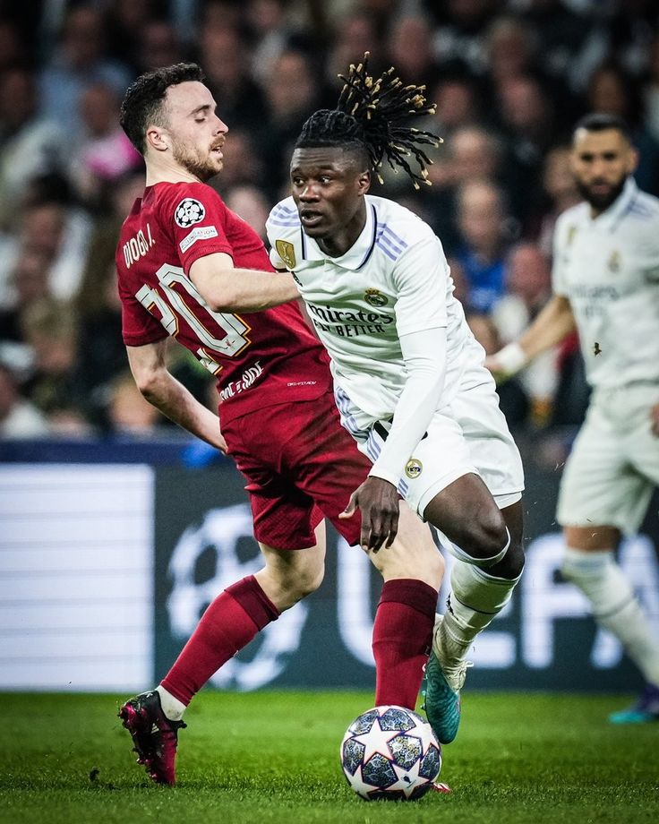 two men playing soccer on a field with people watching from the stands in the background