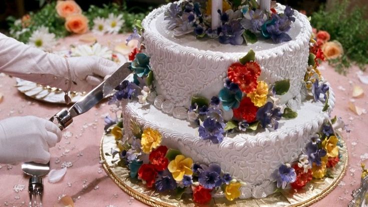 two tiered wedding cake with colorful flowers on the side being cut by a knife