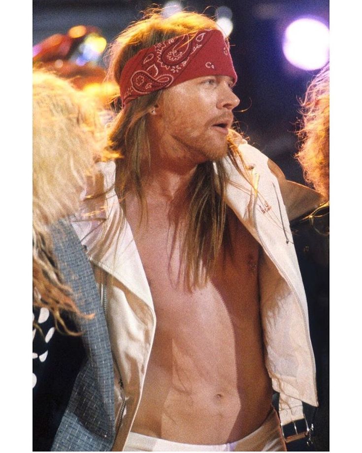 a shirtless man wearing a bandana and looking off into the distance with other people in the background