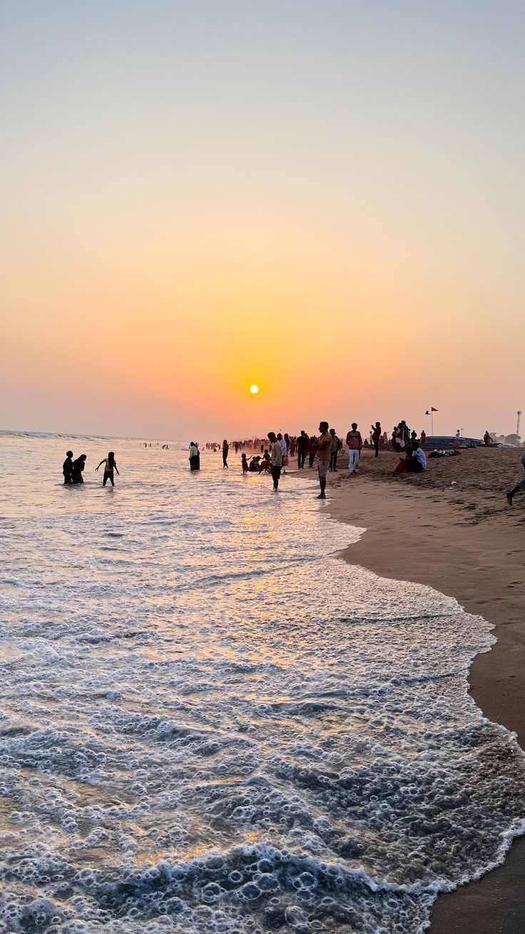 people are on the beach as the sun sets over the water and waves come in