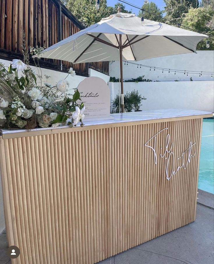 a wooden bar with an umbrella over it next to a swimming pool and some flowers