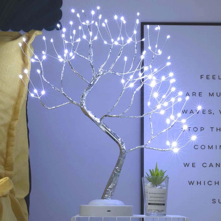 a white tree with lights on it sitting next to a poster and potted plant