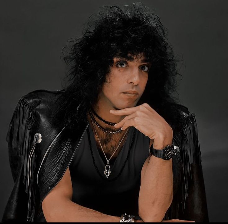 a man with long black hair wearing a leather jacket and bracelets, posing for the camera