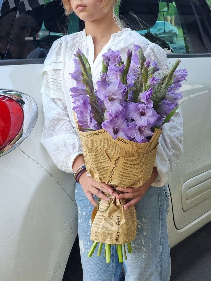 a woman standing next to a car holding a bouquet of purple flowers in her hands