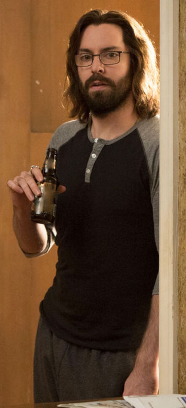 a man with long hair and glasses holding a beer in his hand while standing next to a wooden door