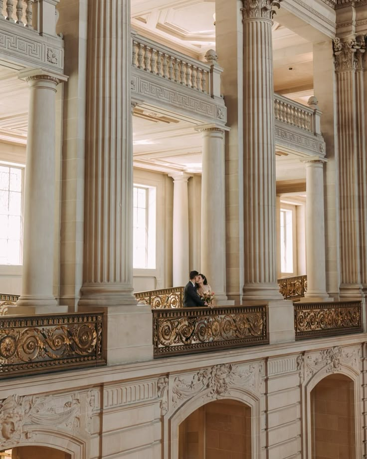 a man and woman standing on the balcony of a large building with columns, pillars and arches