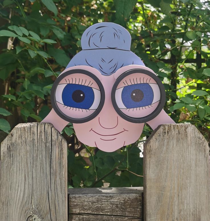 a paper doll with large blue eyes peeking over a wooden fence