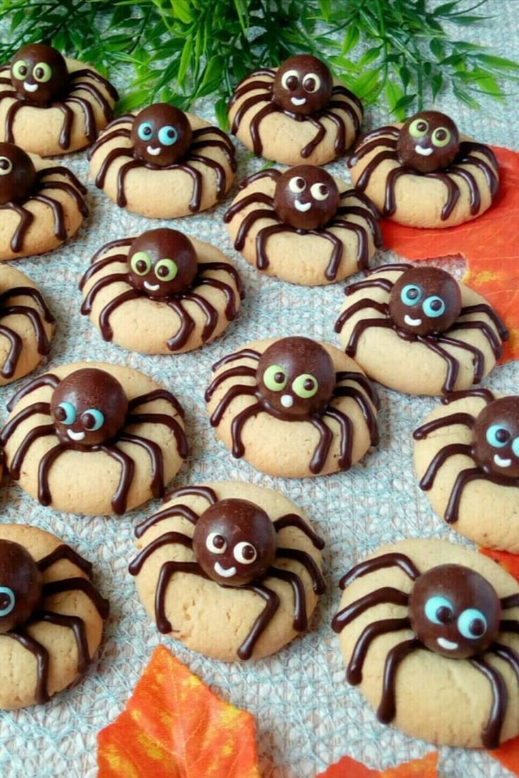 chocolate spider cookies with eyes and googly eyes are arranged on an orange cloth next to green leaves