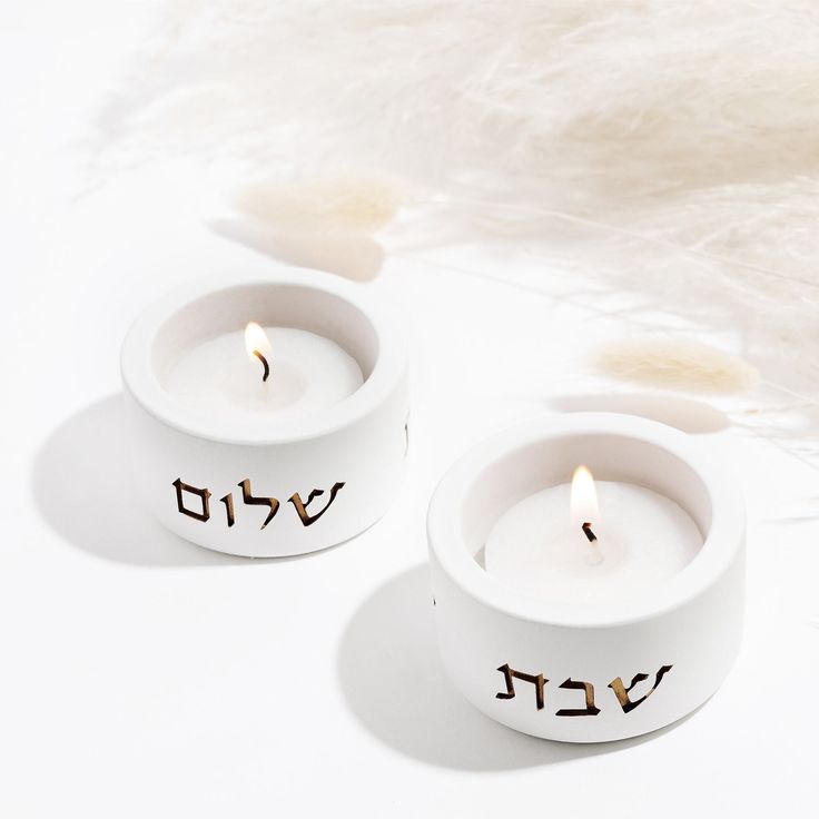 two white candles with hebrew writing on them sitting in front of a fluffy white background