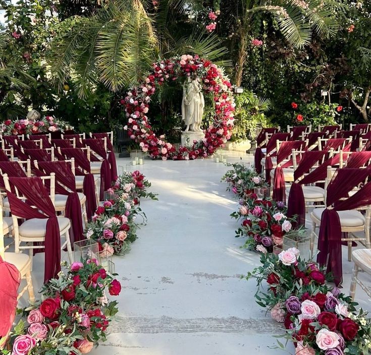 an outdoor ceremony setup with red and pink flowers on the aisle, white chairs and greenery