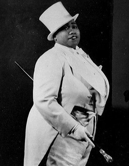 a black and white photo of a woman in a top hat with a cane standing next to a piano