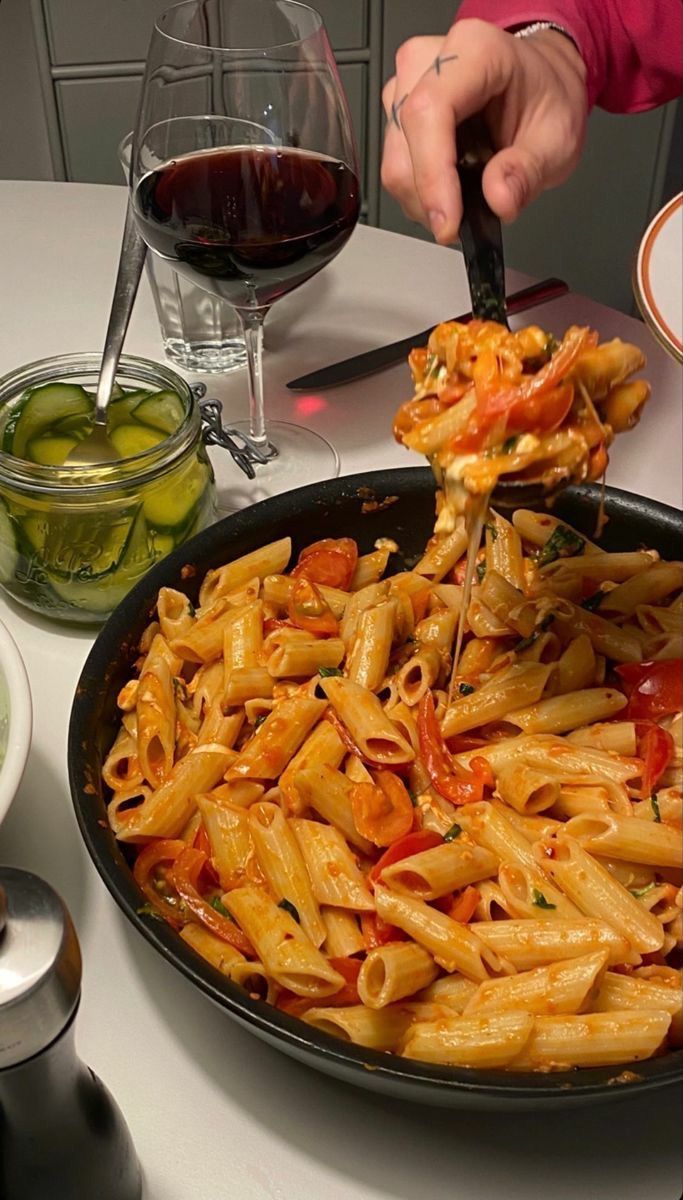 a pan filled with pasta and vegetables on top of a table next to glasses of wine