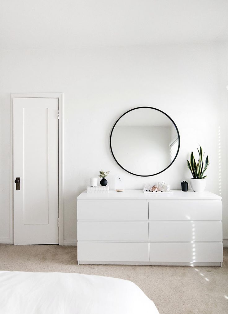 a white bedroom with a round mirror on the wall and dresser in front of it