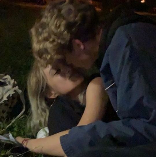 a man and woman are kissing in the grass at night with lights on behind them