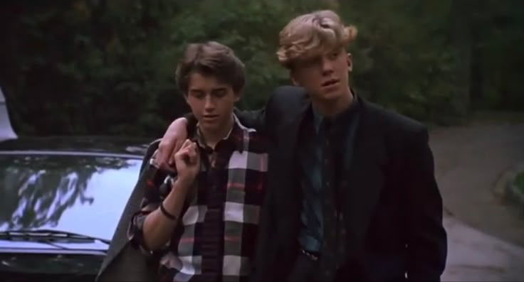 two young men standing next to each other in front of a parked car and looking at each other