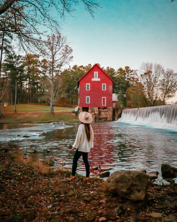 a woman standing in front of a red barn next to a river with waterfall and water fall