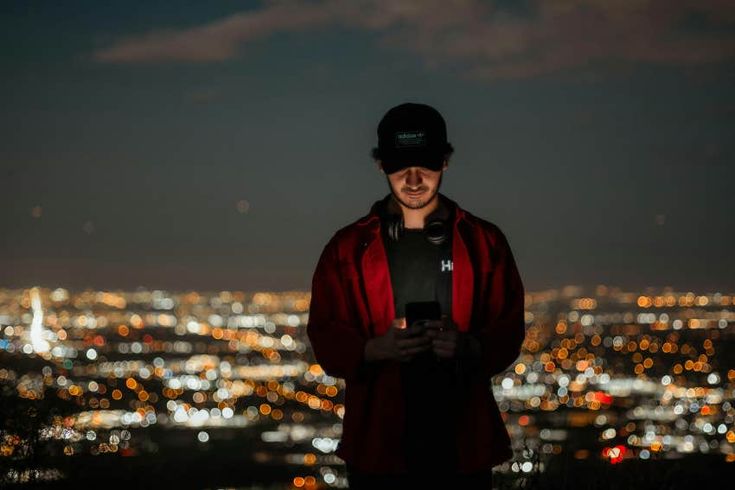 a man standing in front of a city at night looking at his cell phone while wearing a hat