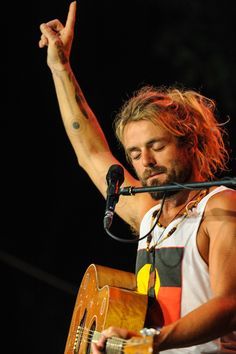 a man with long hair holding a guitar in front of a microphone and giving the peace sign