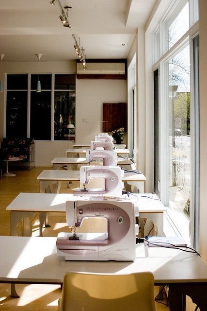 rows of sewing machines lined up on tables in a room with large windows and sun shining through them