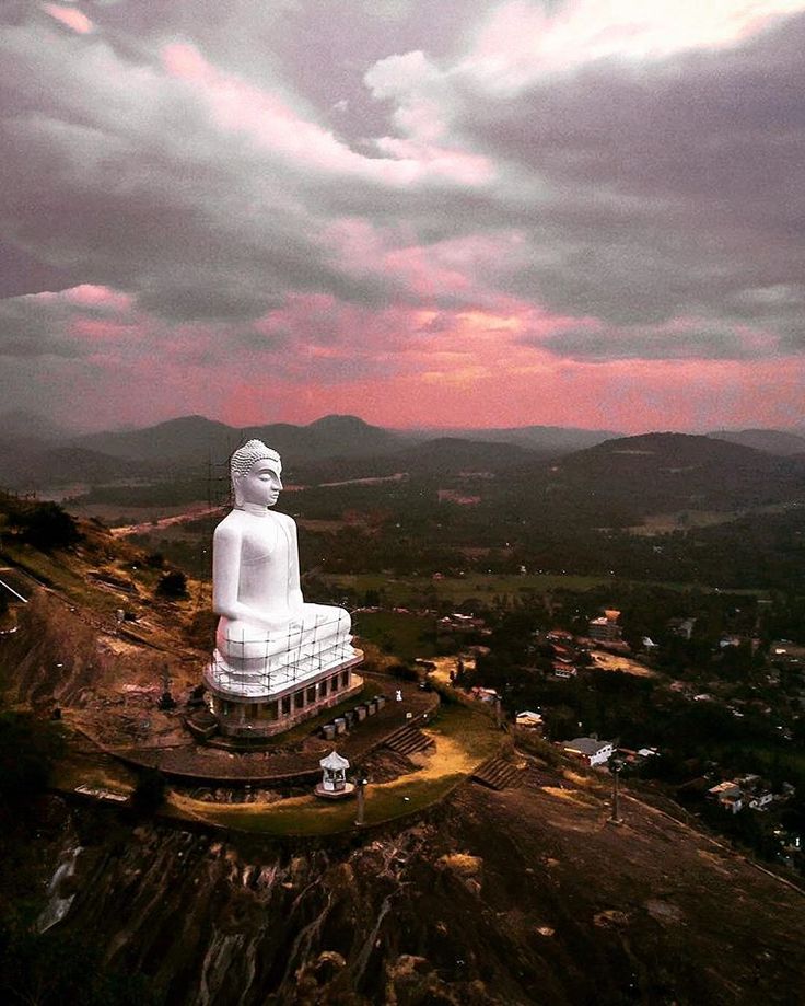 a large white buddha statue sitting on top of a lush green hillside under a cloudy sky