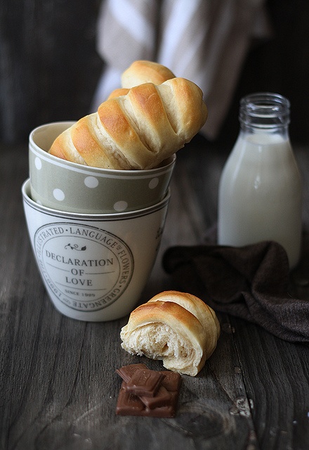 two cups filled with bread next to some milk and chocolate pieces on a table in front of them