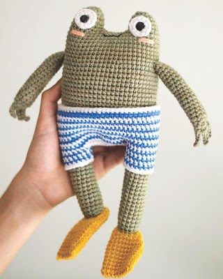 a hand holding a crocheted stuffed animal with one eye open and two legs crossed