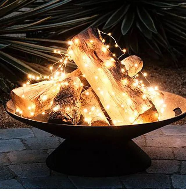 an outdoor fire pit with christmas lights on the top and logs in the bottom, surrounded by plants