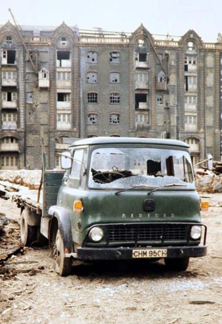 an old green truck is parked in front of a large building with broken down windows