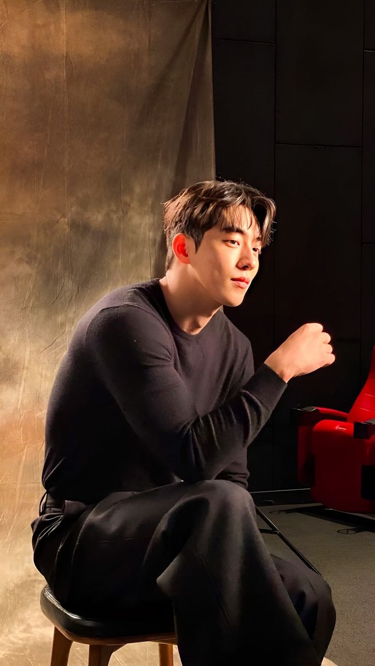 a young man sitting on top of a wooden stool in front of a red chair