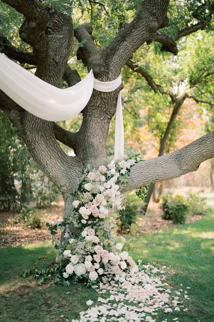 an outdoor wedding ceremony with white flowers and greenery on the ground, in front of a large tree