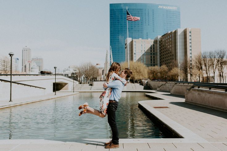 two people standing on the edge of a body of water with buildings in the background