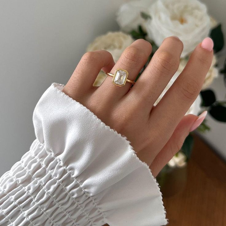 a woman's hand with a gold ring on top of her finger and flowers in the background