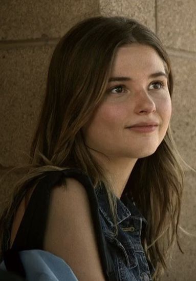 a close up of a person wearing a denim shirt and jeans vest sitting against a brick wall