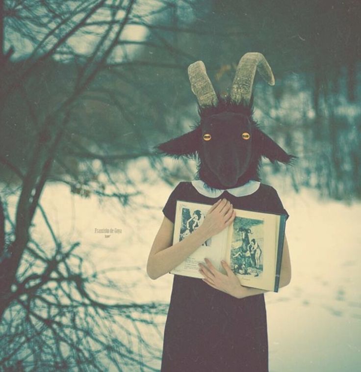 a woman holding an open book with a goat mask on her head, standing in the snow