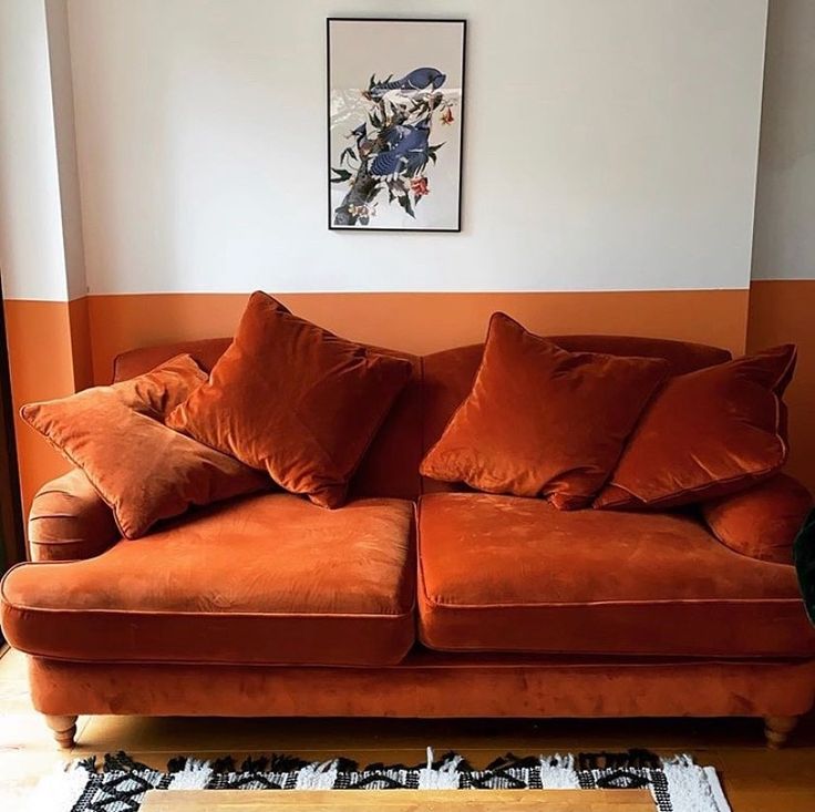 an orange couch with pillows on it in front of a window and rugs against the wall