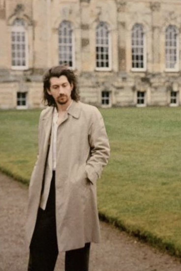 a man with long hair standing in front of a large building wearing a trench coat