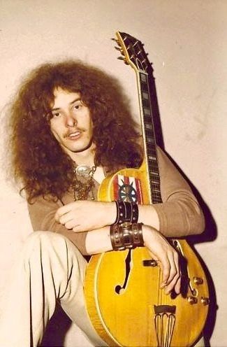 a man with long hair sitting on the ground holding an electric guitar in his hands