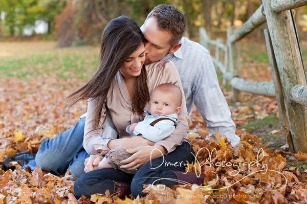 a man and woman holding a baby while sitting on leaves in front of a fence