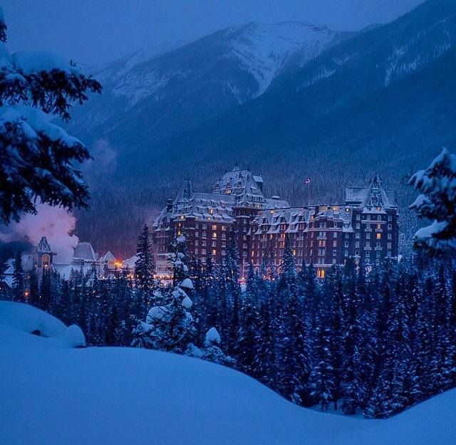 a large building in the middle of a snowy forest with mountains in the background at night