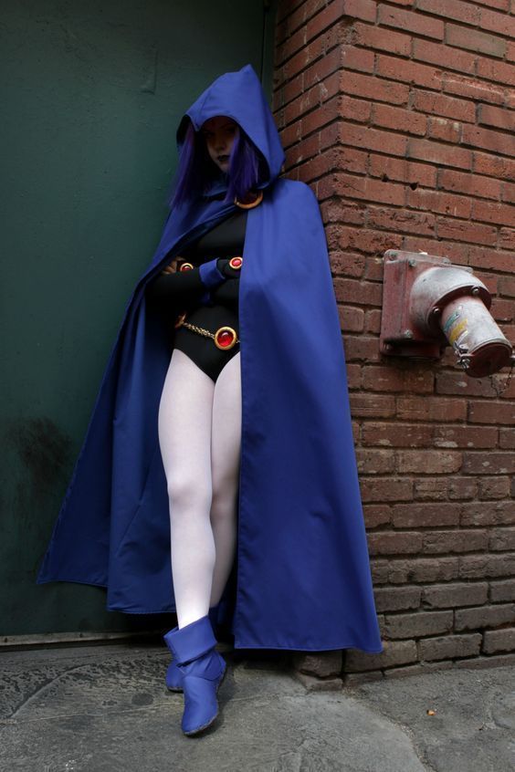 a woman dressed up as a super hero leaning against a brick wall wearing a blue cape and white tights