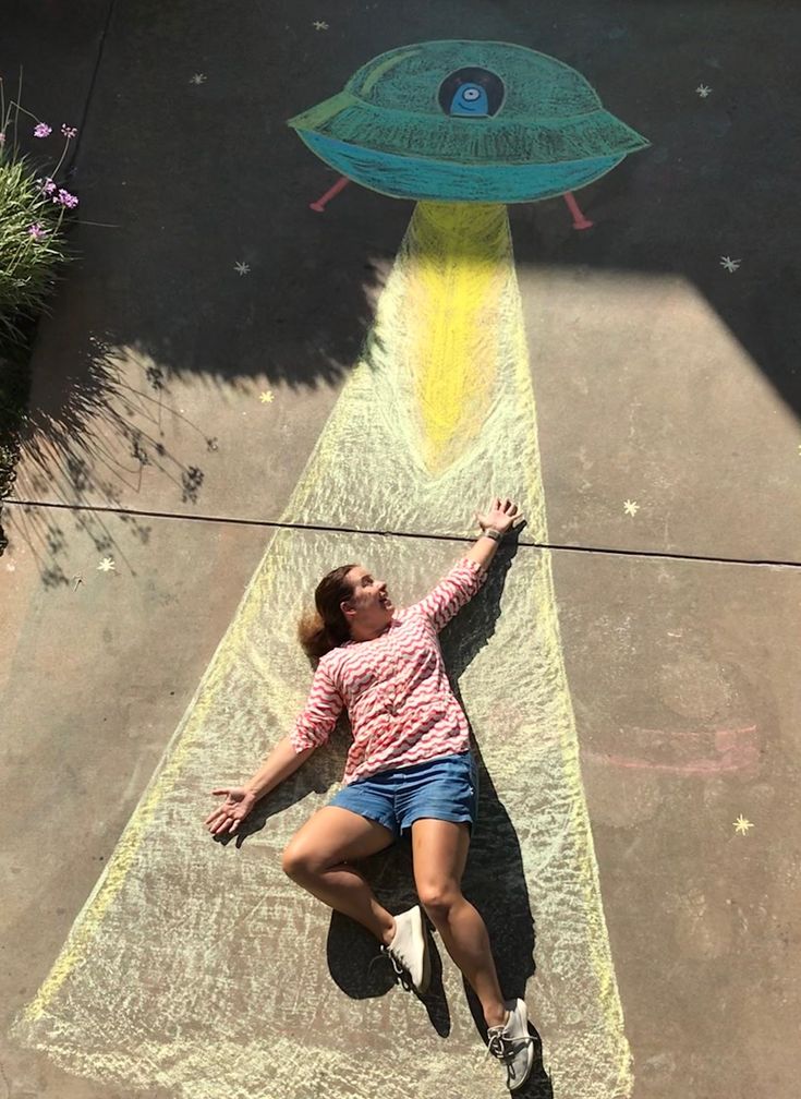 a woman laying on top of a sidewalk next to a chalk drawing alien ship and flying saucer